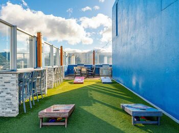 A rooftop area with green turf, cornhole boards, bar seating, and blue walls under a partly cloudy sky.
