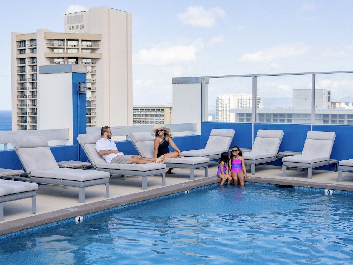 A rooftop pool with people lounging on chairs and sitting by the water, overlooking a city skyline under a clear blue sky.
