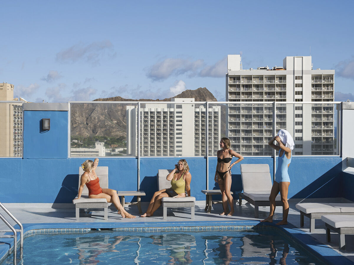 People relaxing by a rooftop pool with city buildings in the background under a clear blue sky.