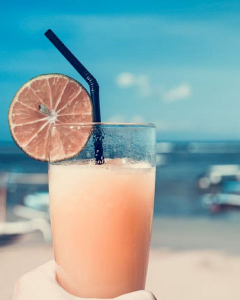 A hand holding a refreshing drink with a lime slice and straw, set against a clear blue sky and beach background.