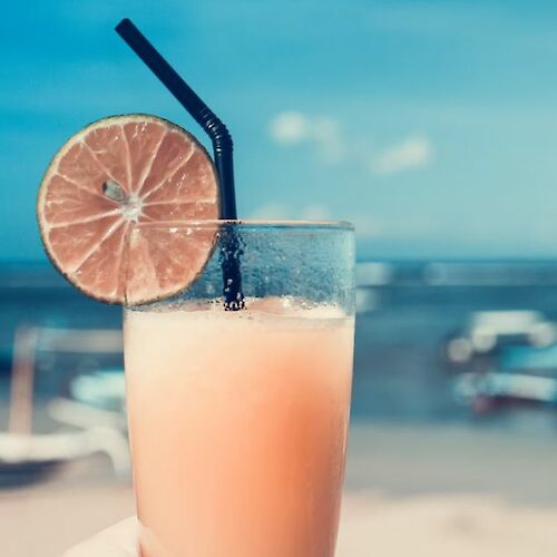 A hand holding a refreshing drink with a lime slice and straw, set against a clear blue sky and beach background.